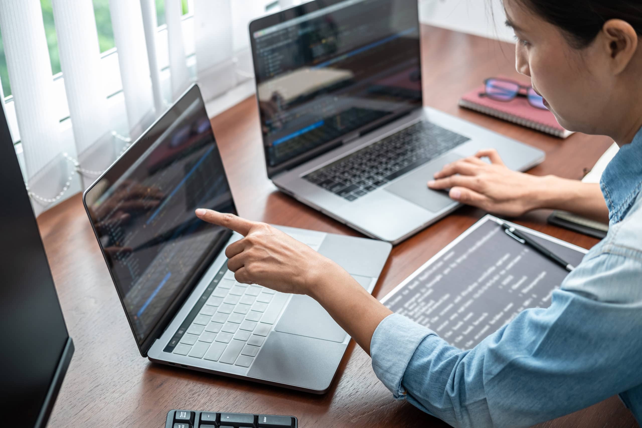 Asian programmer woman pointing and looking on multiple laptop screen to writing code and database while working about development website or applications in software development office