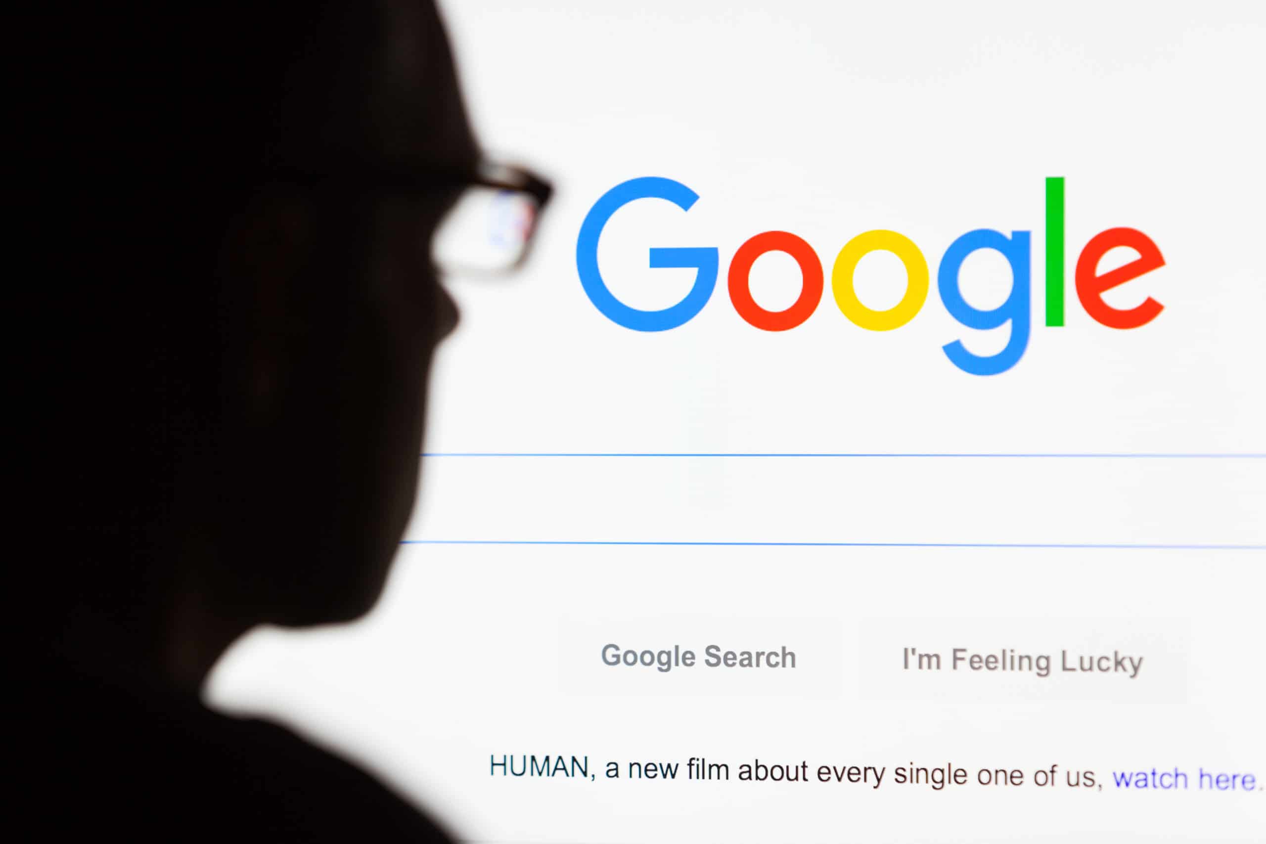 Close-up of the Google.com search homepage displayed on a LCD computer screen with the silhouette of a man's head out of focus in the foreground