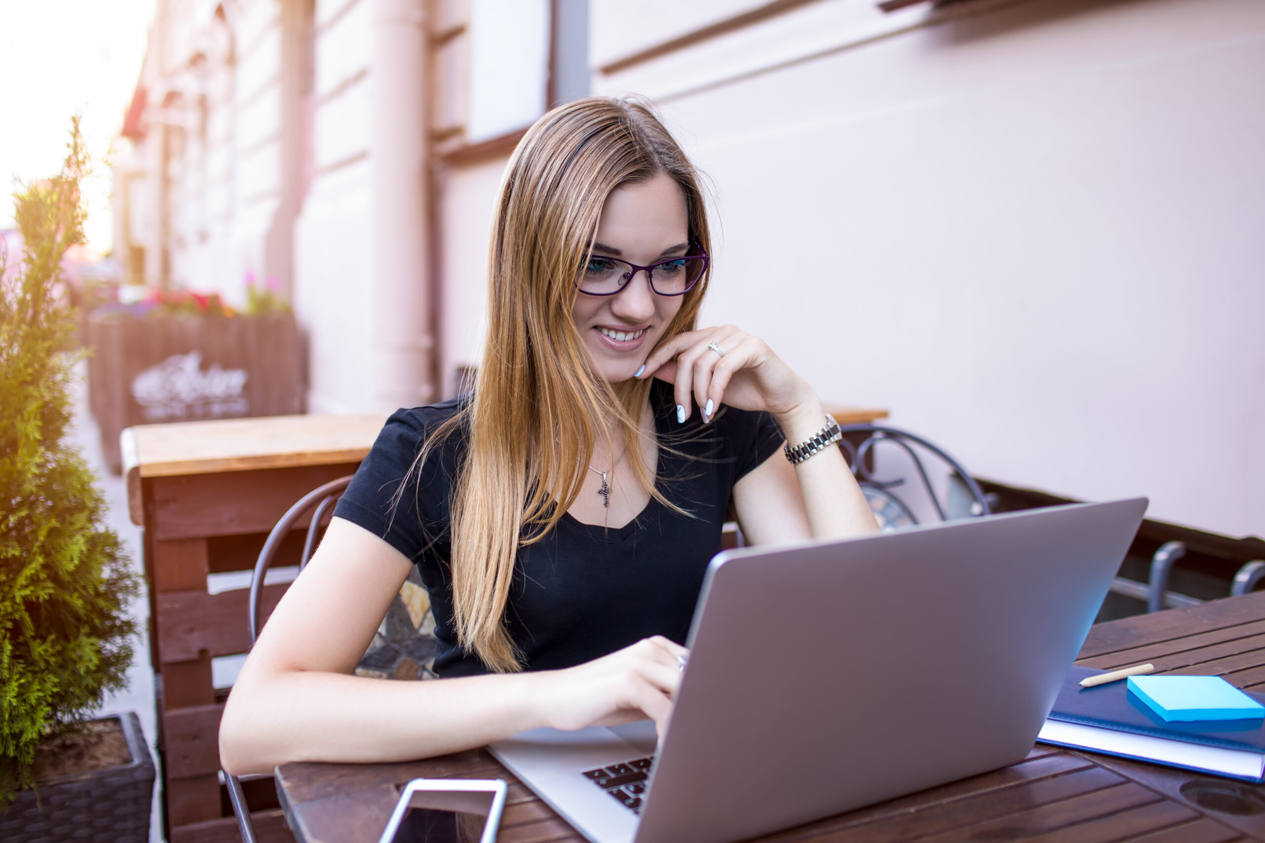 Beautiful smiling woman publication specialist reading article on web site while sitting with laptop computer in street coffee shop.Joyful female university student having video conference via netbook