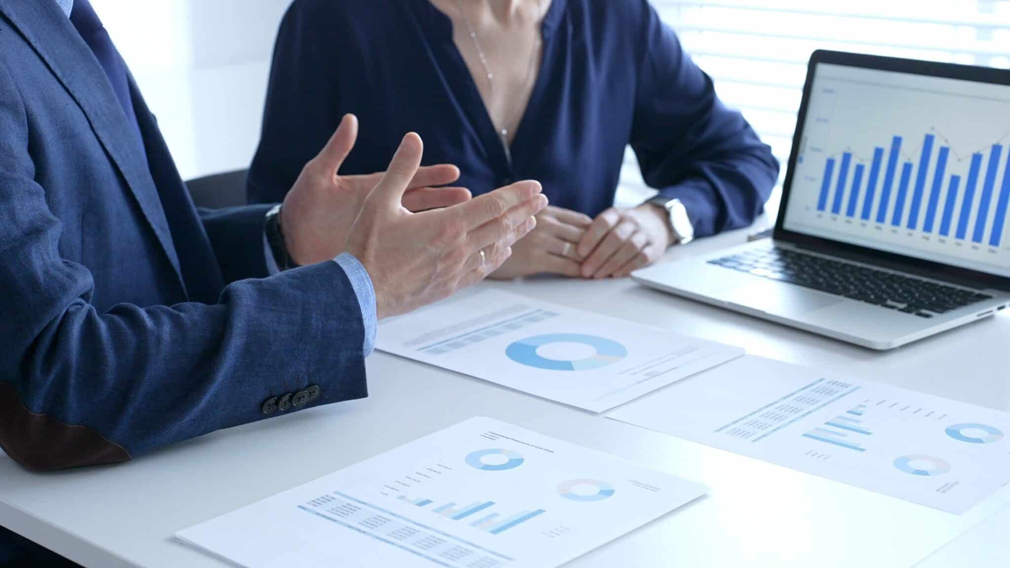 Businessman and business woman reviewing financial documents, collaborating over laptop screen, analyzing market charts for strategic corporate planning. Businesspeople and finance concept.